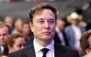 Man in a dark suit with a white shirt and black tie at a formal event, with a crowd in the blurred background.
