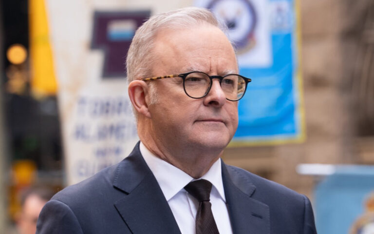 Older man in a dark suit and glasses stands outdoors, looking forward with a neutral expression against a blurred city background.
