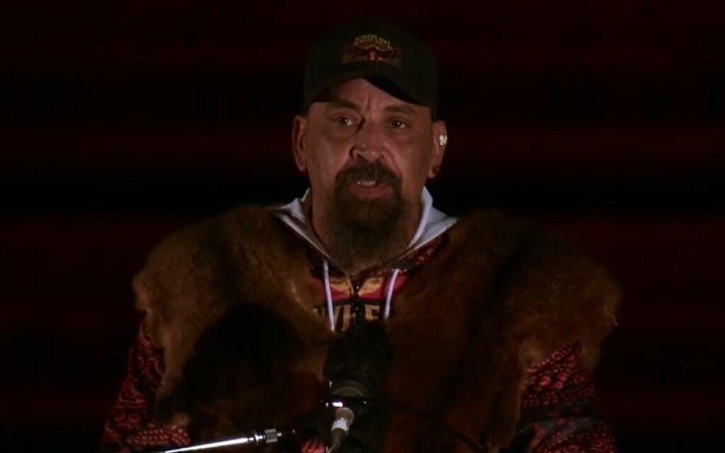 Man wearing a brown fur coat and dark cap speaks into a microphone on a dimly lit stage.