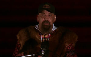 Man wearing a brown fur coat and dark cap speaks into a microphone on a dimly lit stage.