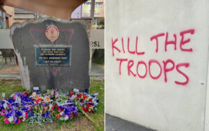 Memorial boulder with a blue plaque and a colorful wreath display in front; plaque text includes a war remembrance message and a crest above it.