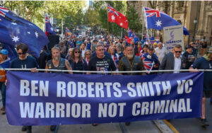 Protesters march down a city street holding a large blue banner that reads 'BEN ROBERTS-SMITH VC WARRIOR NOT CRIMINAL'. Flags fly above the crowd at a rally.