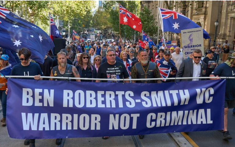 Protesters march down a city street holding a large blue banner that reads 'BEN ROBERTS-SMITH VC WARRIOR NOT CRIMINAL'. Flags fly above the crowd at a rally.