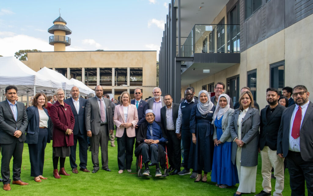 Diverse group of professionals outdoors on a grassy area, centered by an elderly person in a wheelchair, with a modern building behind and white tents nearby.
