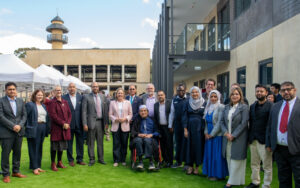 Diverse group of professionals outdoors on a grassy area, centered by an elderly person in a wheelchair, with a modern building behind and white tents nearby.