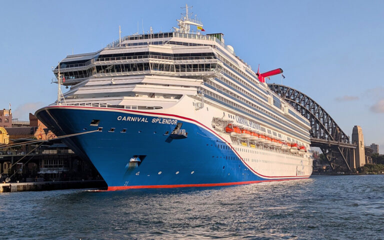 Large Carnival Splendor cruise ship docked at a harbor with a bridge in the background, blue hull and white superstructure.