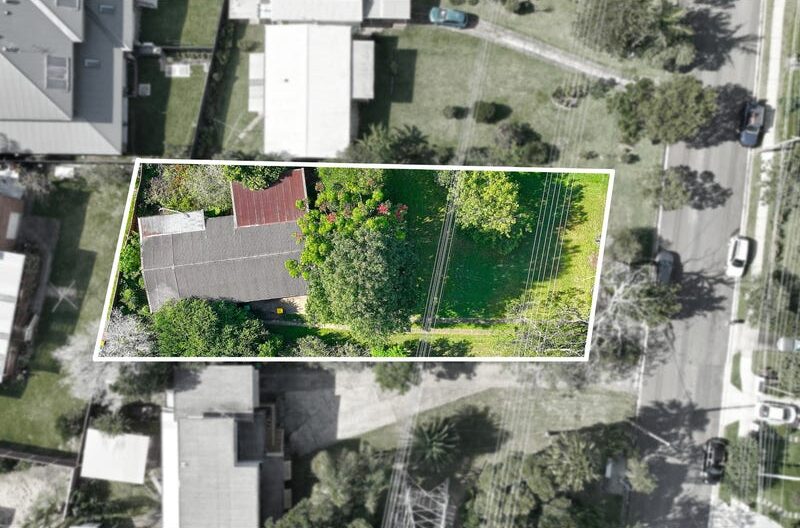 Aerial view of a rectangular residential property outlined in white, showing a house on the left, trees, and a green lawn with power lines along the right edge.