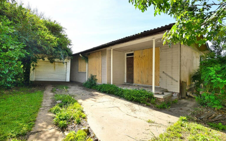 Abandoned single-story house with boarded windows and a detached garage, overgrown yard and cracked driveway within a leafy yard.