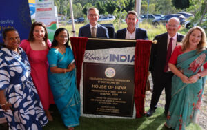 Group of seven people standing beside a commemorative plaque reading 'House of India' during a ceremony outside. (informative)