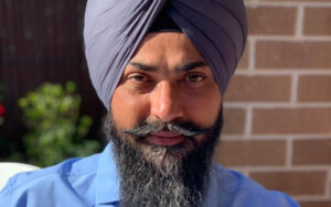 Portrait of a man wearing a gray turban and full beard, smiling at the camera with a brick wall behind him.