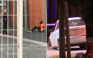 Night scene outside a brick building: a police officer stands near a person seated on the ground behind a chain-link fence.