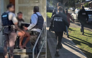 Two officers in tactical vests restrain a seated man on outdoor stairs outside a house, faces blurred or turned away.