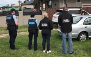 Group of police officers and NDIS staff stand with backs to the camera in a suburban driveway near parked cars outside a house.