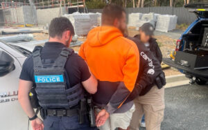 Australian Federal Police officer detaining a man in an orange hoodie at a construction site, with handcuffs visible and another officer nearby.