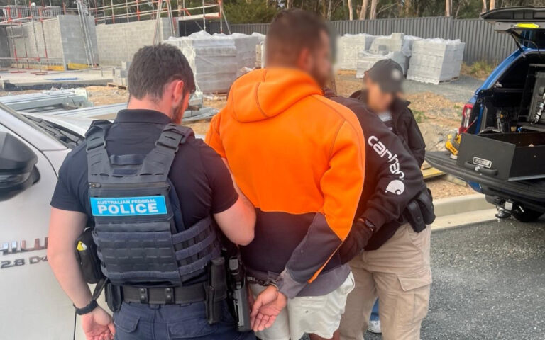 Australian Federal Police officer detaining a man in an orange hoodie at a construction site, with handcuffs visible and another officer nearby.