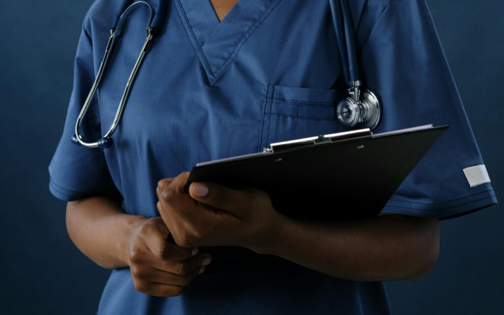 Medical professional in blue scrubs holding a clipboard with a stethoscope around the neck.