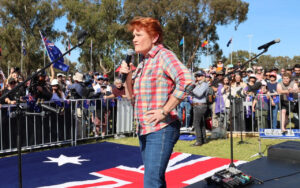 Woman in a plaid shirt speaks into a handheld microphone on an outdoor stage at a rally, with a crowd and Australian flags behind her.