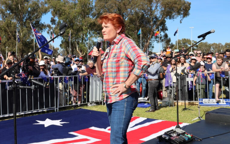 Woman in a plaid shirt speaks into a handheld microphone on an outdoor stage at a rally, with a crowd and Australian flags behind her.