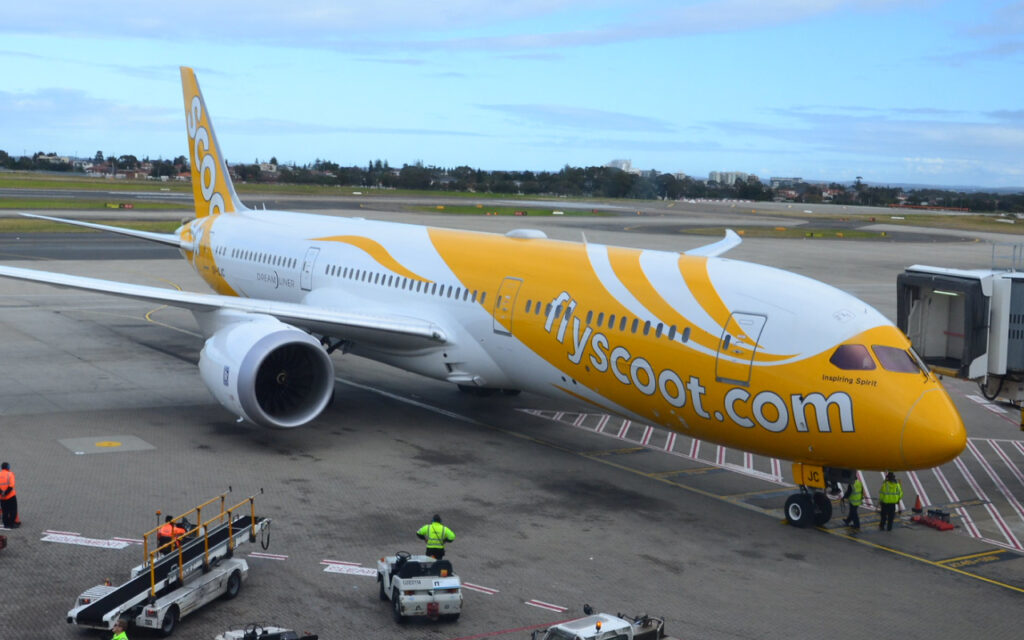 Flyscoot.com airplane parked at a jet bridge on the tarmac with ground crew nearby in safety vests.