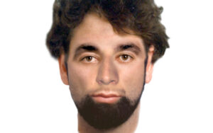 Portrait of a young man with dark hair and a full beard, facing the camera against a white background.