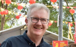 Smiling man with gray hair and glasses at an outdoor festival with red lanterns in the background.