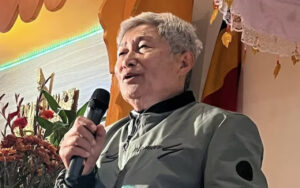 Older man with gray hair speaks into a handheld microphone on a decorated stage, flowers in the background.