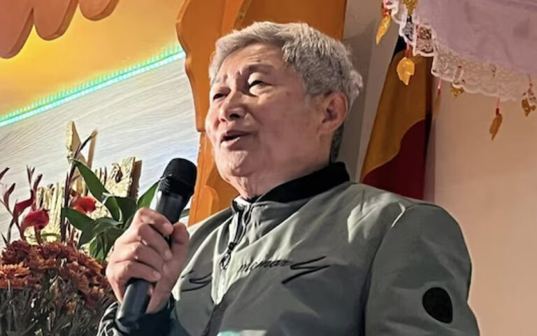 Older man with gray hair speaks into a handheld microphone on a decorated stage, flowers in the background.