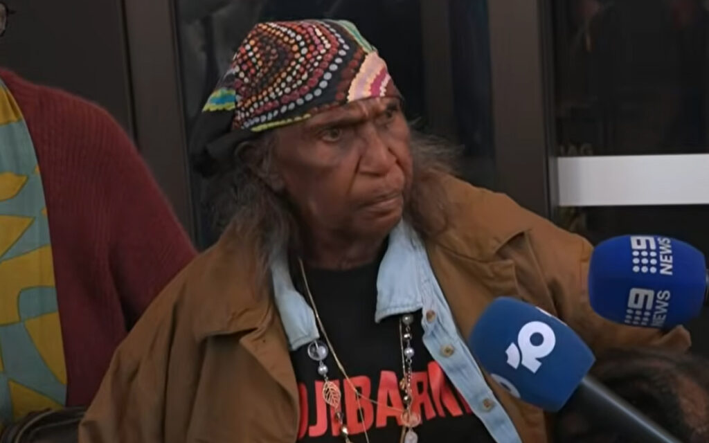 Older man with a colorful beaded bandana and brown jacket speaks to reporters outside, multiple blue mic flags in front of him