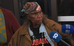 Older man with a colorful beaded bandana and brown jacket speaks to reporters outside, multiple blue mic flags in front of him