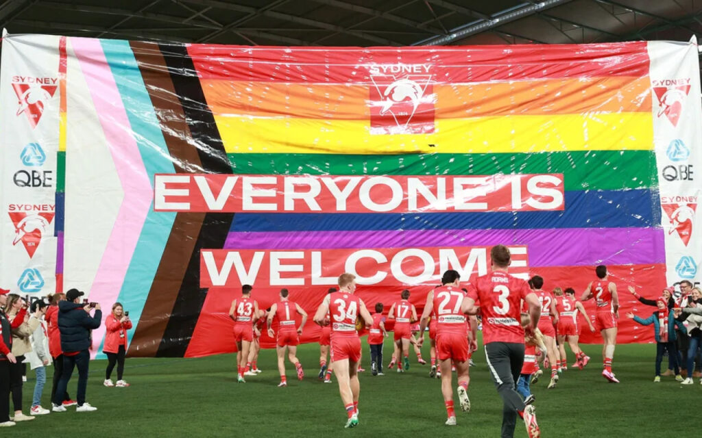 Group of red-uniform athletes running onto a green field under a giant rainbow banner that reads 'EVERYONE IS WELCOME'.