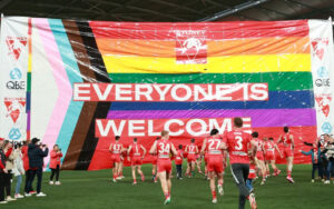 Group of red-uniform athletes running onto a green field under a giant rainbow banner that reads 'EVERYONE IS WELCOME'.