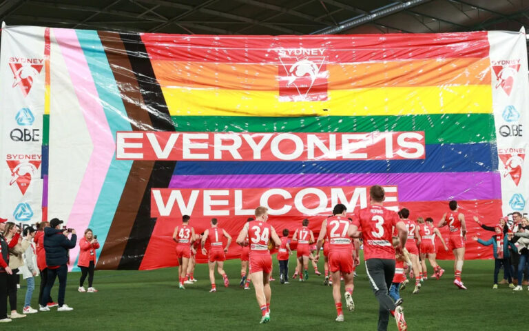 Group of red-uniform athletes running onto a green field under a giant rainbow banner that reads 'EVERYONE IS WELCOME'.