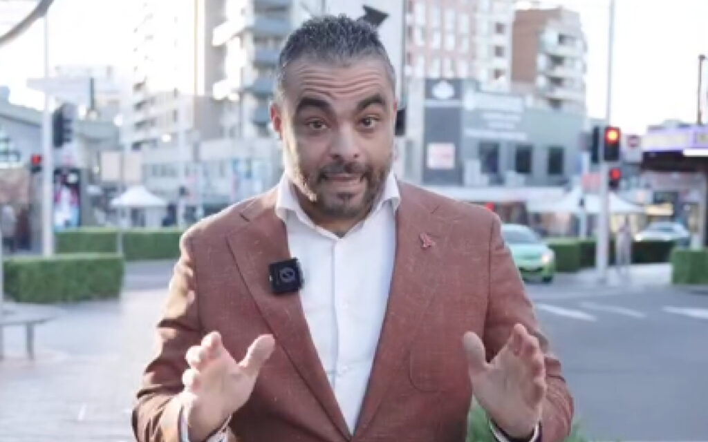 Man in a brown blazer with a lapel mic speaking to the camera on a city street backdrop