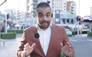 Man in a brown blazer with a lapel mic speaking to the camera on a city street backdrop