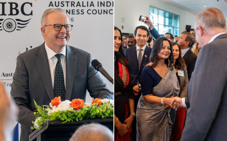 Smiling man in a suit speaks at a podium with orange and white flowers during a formal event; backdrop reads Australian business councilLike text indicating a business gathering.
