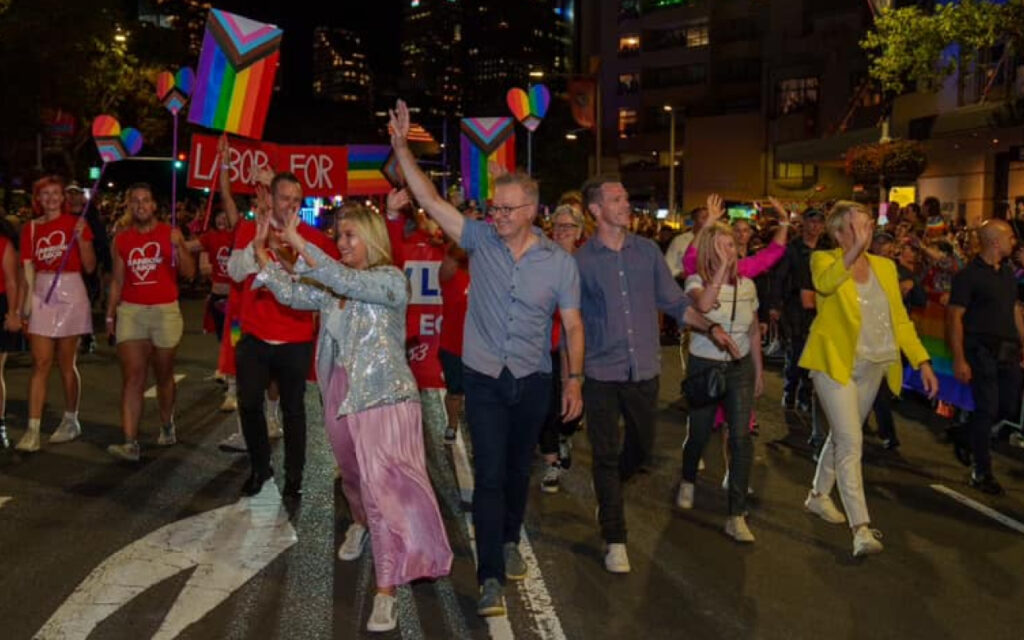 A diverse group of people march in a pride parade at night, carrying rainbow flags and colorful signs down a city street.