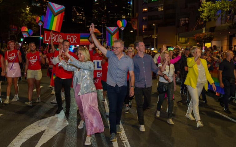A diverse group of people march in a pride parade at night, carrying rainbow flags and colorful signs down a city street.