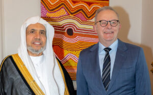 Two men posing indoors in front of a colorful abstract painting: one in traditional arabic robe with white headscarf, the other in a blue suit and striped tie smiling.