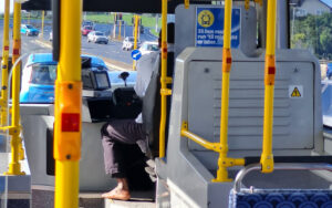 Driver seated at the controls of a bus, with bright yellow handrails and poles surrounding the cabin; cars and a road scene are visible through the windshield.