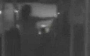 Dim grayscale photo of a dark hallway with a bright rectangular doorway in the center and indistinct shapes nearby.