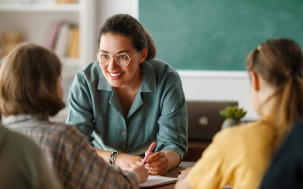 Teacher with glasses leans over a table, smiling, aiding students in a classroom setting with pens and papers on the table.