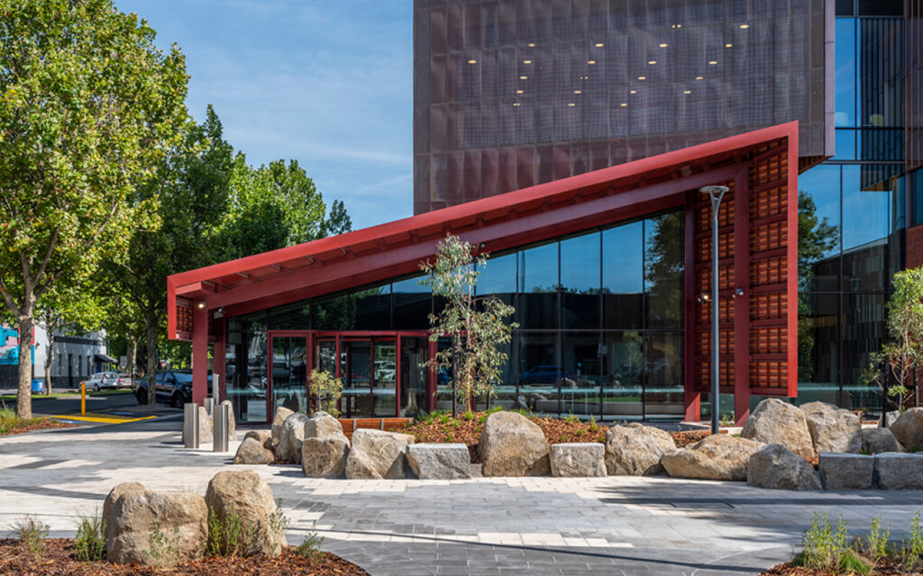 Modern building exterior with a red angled entrance canopy, glass walls, and landscaped rocks and trees in the foreground.