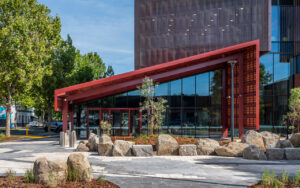 Modern building exterior with a red angled entrance canopy, glass walls, and landscaped rocks and trees in the foreground.