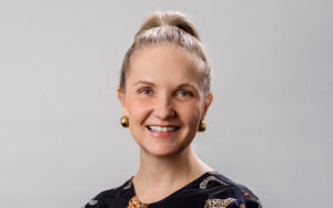 Smiling woman with a blonde hair bun, gold earrings, and a black patterned top against a gray background (head-and-shoulders portrait).","Portrait of a cheerful woman with blue eyes, gold earrings, and a dark patterned blouse.","Head-and-shoulders portrait of a friendly woman in a dark patterned top and gold earrings, against a light gray backdrop.","Close-up portrait of a smiling woman with a high bun, gold earrings, and a black shirt.","Professional headshot of a woman with a ponytail bun, gold earrings, and a warm smile.