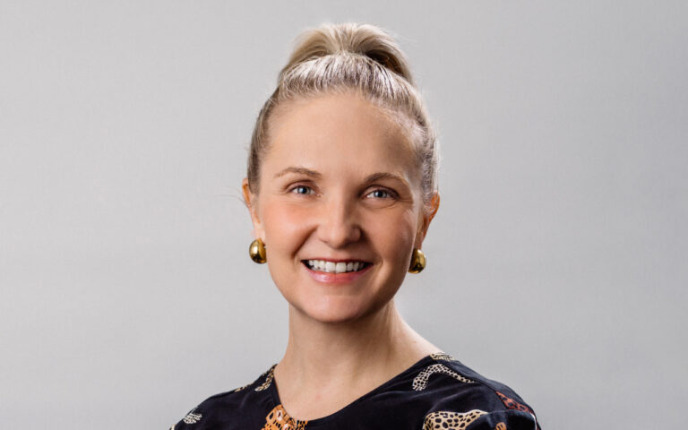 Smiling woman with a blonde hair bun, gold earrings, and a black patterned top against a gray background (head-and-shoulders portrait).","Portrait of a cheerful woman with blue eyes, gold earrings, and a dark patterned blouse.","Head-and-shoulders portrait of a friendly woman in a dark patterned top and gold earrings, against a light gray backdrop.","Close-up portrait of a smiling woman with a high bun, gold earrings, and a black shirt.","Professional headshot of a woman with a ponytail bun, gold earrings, and a warm smile.