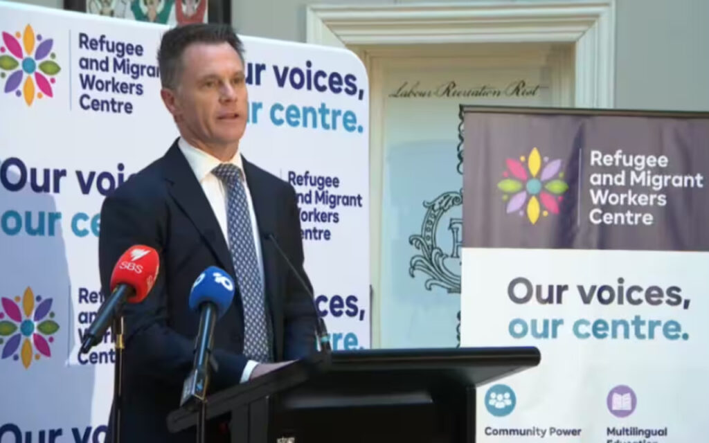 Man in a suit giving a speech at a podium, flanked by banners for the Refugee and Migrant Workers Centre and microphones from multiple news outlets.