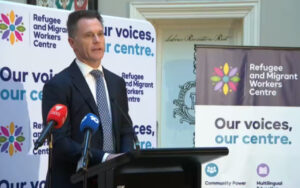 Man in a suit giving a speech at a podium, flanked by banners for the Refugee and Migrant Workers Centre and microphones from multiple news outlets.