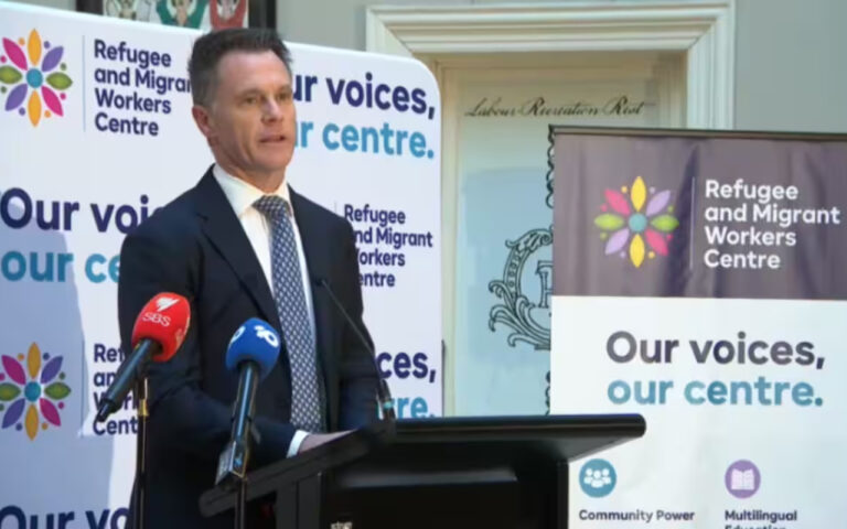 Man in a suit giving a speech at a podium, flanked by banners for the Refugee and Migrant Workers Centre and microphones from multiple news outlets.