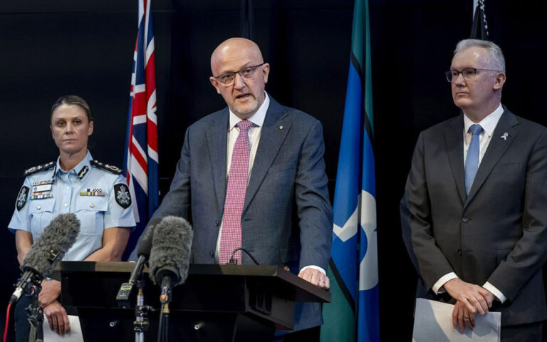 A man in a gray suit and pink tie speaks at a podium flanked by a police officer on the left and a suited man on the right, with microphones in front and flags in the background.