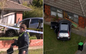 A silver car crashes into a brick house, with police tape marking off the scene and a person in uniform nearby.